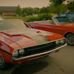 Cherry Red Dodge Challenger and Reddish Orange Ford Mustang sit side by side in front of a water fountain at the September 26th, 2021 Cars & Coffee event in Fitchburg, Wisconsin.
